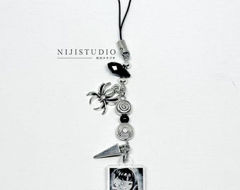 May include: A silver charm with a black cord features a variety of decorative elements. These include a spider, spiral beads, a black gem, and a small square pendant with a black and white image of a person. The text "NIJISTUDIO" is also visible.