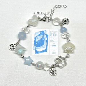 May include: A silver bracelet with a variety of charms, including a heart, star, shell, and spiral designs. The bracelet features blue and clear beads, with a clasp closure. A blue and white graphic is in the background.