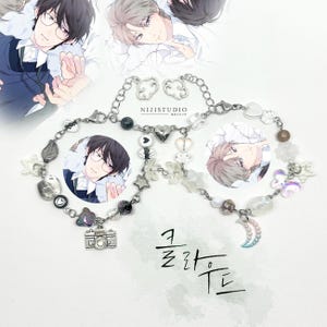 Puede incluir: Dos pulseras de cuentas con abalorios, incluyendo una cámara, estrellas y una luna. Las pulseras presentan cuentas transparentes, blancas y negras, con una cadena plateada. El fondo incluye ilustraciones de personajes de anime y el texto "NIJISTUDIO".
