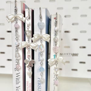 Puede incluir: Cuatro marcapáginas de metal plateado con lazos blancos, cuentas transparentes y de colores pastel, y dijes en forma de corazones y mariposas. Cada marcapáginas cuelga de un clip metálico en el lomo de un libro. Se observa el texto NIJISTUDIO.