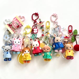 Puede incluir: Una colección de llaveros coloridos con figuras de animales en miniatura, incluyendo un koala, un ratón, un conejo, un gato y un perro, cada uno con un atuendo de diferente color. Los llaveros tienen varios dijes y acentos en forma de fruta.