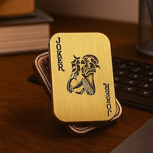 以下が含まれることがあります： 金色の金属製ジョーカーカード。カードの両側に「JOKER」の文字と、ジョーカーの人物の精巧なイラストが描かれています。カードは、同様の濃い色のカードの上に置かれています。