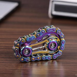 Peut inclure: Un fidget spinner en métal multicolore avec un design de chaîne de vélo. Le spinner présente une finition irisée arc-en-ciel avec des accents dorés et violets. La conception comprend deux composants circulaires et un axe central.
