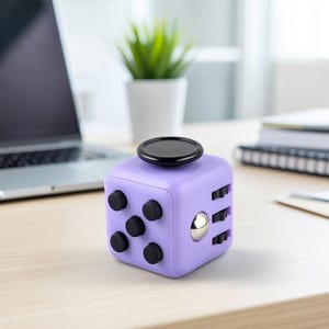 Könnte beinhalten: Ein lila Fidget Cube mit schwarzen Knöpfen, einer silbernen Kugel und einer schwarzen Scheibe oben. Der Würfel steht auf einer hellen Holzoberfläche, im Hintergrund sind ein Laptop und eine kleine Topfpflanze zu sehen.
