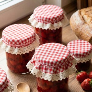 Peut inclure: Plusieurs pots de confiture de fraises faits maison avec des couvercles à carreaux rouges et blancs et une bordure en dentelle. Une cuillère en bois et des fraises fraîches sont posées sur une surface en bois, à côté d'un pain.