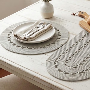 May include: A dining table setting with a white plate, silverware, and a folded linen napkin on a gray placemat. A matching oval runner is next to the plate. A small vase with greenery and a wooden cutting board with bread and cheese are also on the table.