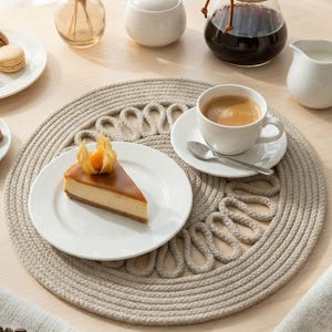 May include: A coffee setup featuring a slice of cheesecake on a white plate, a glass coffee maker, a cup of coffee, and a small milk jug. A beige placemat and a vase with dried flowers are also visible, creating a cozy atmosphere.