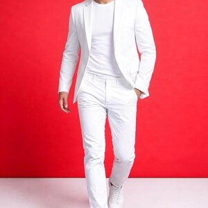 May include: A white suit, consisting of a blazer and trousers, paired with a white t-shirt and sneakers. The outfit is set against a vibrant red background, creating a striking contrast. The suit appears to be made of a smooth fabric.