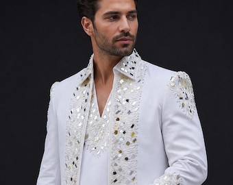 May include: A white blazer and matching shirt set featuring intricate embroidery and reflective mirror embellishments along the lapels, collar, shoulders, and cuffs. The garment displays a structured fit with a V-neckline and long sleeves.