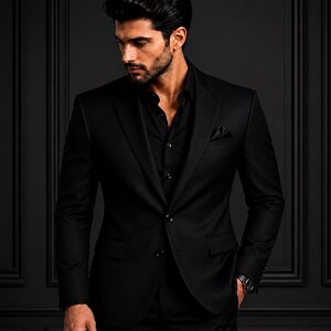 May include: A solid black blazer worn over a matching black button-down shirt. The blazer features a notched lapel, two front buttons, and a pocket square in the breast pocket. A dark wristwatch is visible on the left wrist of the wearer.
