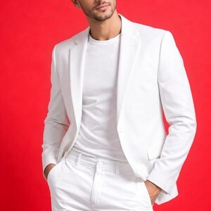 May include: A person wearing a white suit, including a blazer, t-shirt, and pants, against a vibrant red background. The suit is tailored and appears to be made of a smooth fabric. The person has their hands in their pockets.
