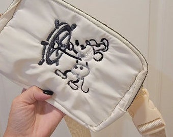 May include: A white canvas crossbody bag with a black embroidered image of Mickey Mouse holding a ship's wheel. The bag has a cream-colored adjustable strap.