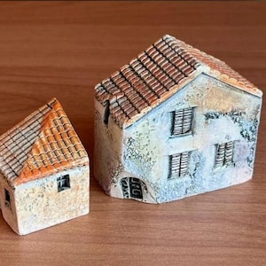 Op de afbeelding: Twee miniatuur keramische huizen met gedetailleerde kenmerken. De huizen hebben oranje-bruine pannendaken en gebroken witte muren met kleine ramen. Het grotere huis heeft een deur en meerdere ramen. De huizen staan op een houten oppervlak.