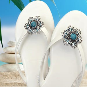 May include: A pair of white flip-flops with silver flower embellishments featuring turquoise stones. The flowers are detailed with intricate designs.