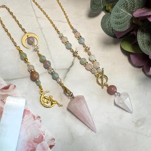 May include: Two gold-toned metal chains featuring assorted pastel-colored beads, star-shaped charms, and a crescent moon charm with a fairy figure. One chain ends in a large, pointed rose quartz pendant, while the other features a clear crystal point.