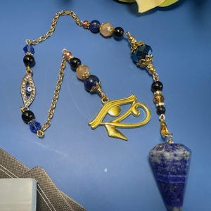 May include: A gold-toned chain pendulum featuring a faceted blue lapis lazuli stone point, a gold-toned Eye of Horus charm, and a smaller evil eye charm. The chain is decorated with various beads in shades of blue, black, and beige, with gold-toned metal accents.