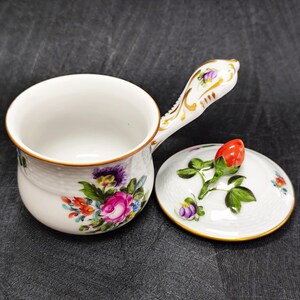 Könnte beinhalten: Eine weiße Porzellansaucenpfanne mit floralem Design und goldenem Rand. Der gebogene Griff ist mit goldenen Akzenten verziert. Der Deckel zeigt eine rote Rosenknospe und grüne Blätter. Der Topf hat einen Durchmesser von ca. 10 cm.