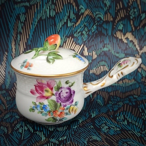 Puede incluir: Una salsera de cerámica blanca con tapa, con un diseño floral en rosa, morado, azul y naranja. La olla tiene un borde y un asa dorados, y una fruta decorativa en la tapa. La olla tiene aproximadamente 15 cm de diámetro.