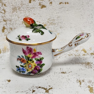 Puede incluir: Cacerola de cerámica blanca con tapa y asa, adornada con un diseño floral. La olla presenta un borde y detalles de asa dorados. Un capullo floral rojo y verde decora la tapa. La olla está decorada con flores coloridas.