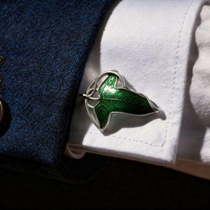 Puede incluir: Gemelos plateados con un diseño de hoja verde. La hoja presenta vetas detalladas y un acabado brillante. Los gemelos se colocan sobre un puño de camisa blanco y una blazer azul marino, destacando el estilo elegante del accesorio.
