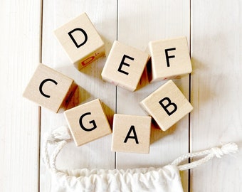 May include: Wooden alphabet blocks with the letters C, D, E, F, G, A, and B painted in black on each block. The blocks are arranged in a semi-circle on a white wooden surface. The blocks are in a white drawstring bag with the words "Musical Alphabet Blocks" printed on the bag.