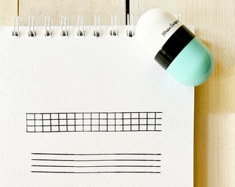 May include: A white and blue capsule-shaped eraser with the text "Mouse Coop" on the side. The eraser is sitting on a white notebook with a black grid and lines drawn on the page.