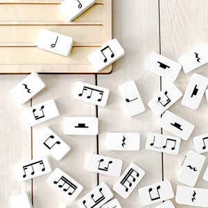 May include: White rectangular blocks with black musical notes and symbols scattered on a light wood surface. The blocks feature various musical notations, including quarter notes, eighth notes, and rests. A wooden tray is visible in the background.
