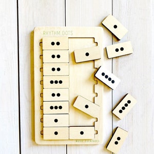 May include: Wooden rhythm dots game with a board and separate tiles. The board has a label that says "Rhythm Dots" and "Music Escapades". The tiles have black dots on them, ranging from one to four dots.