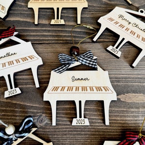 May include: A set of wooden piano ornaments with black and white checked ribbon bows. Each ornament is personalised with a name and the year 2023. The ornaments are perfect for a Christmas tree or as a gift for a music lover.