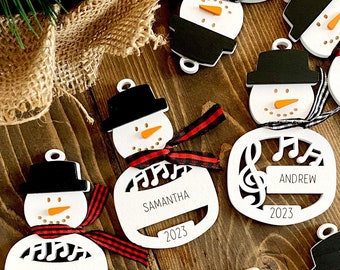 May include: White wooden snowman ornaments with black hats and scarves. Each ornament has a musical note design and a personalized name, including "Samantha" and "Andrew". The ornaments also include the year "2023".
