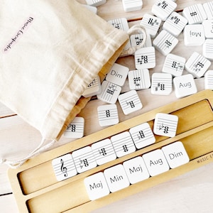 May include: A music learning set with white tiles featuring musical symbols and terms like "Maj" and "Dim." A canvas bag with "Music Escapades" printed on it is next to the tiles. A wooden tray holds some of the tiles.