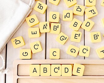 May include: A set of yellow wooden tiles with letters and musical sharps and flats. The tiles are arranged in a row on a wooden board, spelling out the musical notes A, B, C#, D, and E.