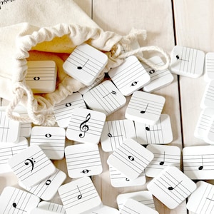 May include: A white canvas bag filled with white wooden tiles. Each tile has a different musical note or symbol printed on it. The tiles are scattered on a light wood surface.