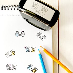 May include: A white paper with four black and white hand outlines with numbers 1-5 on each finger. The hands are labeled "LH" and "RH" for left and right hand. The paper is on a wooden surface with two colored pencils, one blue and one orange.