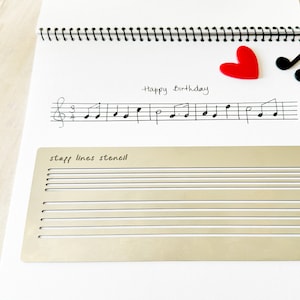 May include: A white sheet of paper with a handwritten message that says "Happy Birthday". There is a musical staff with notes drawn on it. A red heart and a treble clef are drawn on the top of the page. A gold metal stencil with the words "staff lines stencil" is on the bottom of the page.