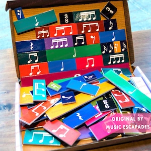 May include: A colorful set of magnetic music tiles featuring musical notes, clefs, and numbers. The tiles are arranged in a box with the words "ORIGINAL BY MUSIC ESCAPADES" printed on the bottom.