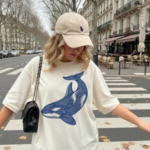 May include: Cream-colored t-shirt featuring a blue and white orca design. The shirt is paired with a tan baseball cap with a small navy blue logo. A black shoulder bag with a chain strap completes the look.
