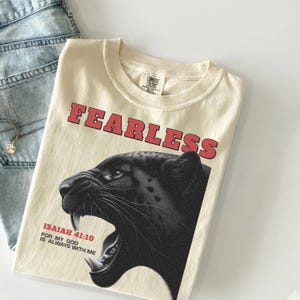 May include: Cream-coloured t-shirt featuring the word "FEARLESS" in red block letters. A black panther graphic is below the text. The shirt also includes the text "ISAIAH 41:10 FOR MY GOD IS ALWAYS WITH ME."