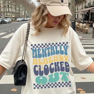 May include: A cream-colored t-shirt featuring a graphic with the text "MENTALLY ALREADY CLOCKED OUT" in wavy, multi-colored letters. The design includes a checkered border at the top and bottom, accented with small yellow star shapes throughout.