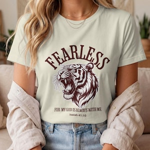 May include: A cream-coloured t-shirt featuring the word "FEARLESS" in maroon above a detailed illustration of a roaring tiger. Below the tiger is the text "FOR MY GOD IS ALWAYS WITH ME" and "Isaiah 41:10" in maroon.