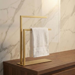 Pode incluir: Um toalheiro de metal dourado com base retangular e estrutura vertical. Uma toalha branca está pendurada no toalheiro. O toalheiro está sobre uma superfície de madeira marrom escura, com uma parede com padrão de mármore ao fundo.