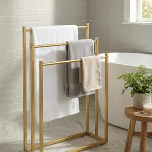 Pode incluir: Um toalheiro de metal dourado com três toalhas num ambiente de casa de banho. As toalhas são brancas, cinzentas e bege. O toalheiro tem uma base retangular e três barras horizontais para pendurar toalhas. A casa de banho tem uma banheira branca e um pequeno banco de madeira.