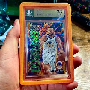 May include: A basketball trading card of Stephen Curry, graded 9.5 by Beckett, in an orange protective case. The card features Curry in his Golden State Warriors uniform, holding a basketball. The card's design includes the text "EN FUEGO".