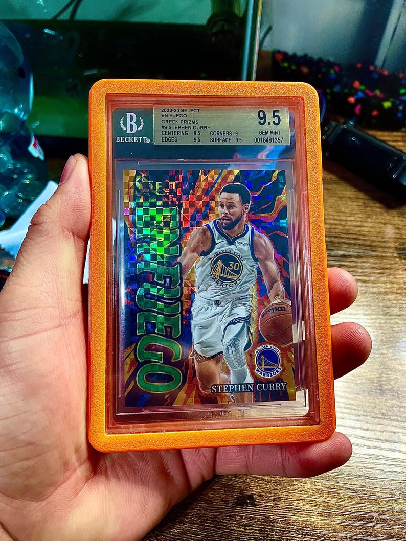 May include: A basketball trading card of Stephen Curry, graded 9.5 by Beckett, in an orange protective case. The card features Curry in his Golden State Warriors uniform, holding a basketball. The card's design includes the text "EN FUEGO".