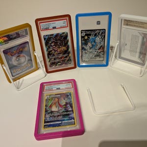 May include: A collection of graded trading cards in protective cases, displayed in clear plastic stands. The cases are various colors, including gold, red, blue, white, and pink. The cards feature colorful artwork and text.