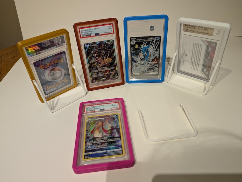 May include: A collection of graded trading cards in protective cases, displayed in clear plastic stands. The cases are various colors, including gold, red, blue, white, and pink. The cards feature colorful artwork and text.
