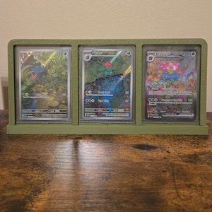 Könnte beinhalten: Ein grüner Display-Ständer mit drei Pokémon-Sammelkarten. Die Karten zeigen Bulbasaur, Ivysaur und Venusaur mit lebendigen Kunstwerken und holografischen Details. Der Ständer besteht aus einem matten, grünen Material.