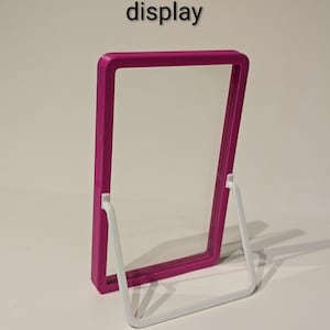 May include: A magenta display frame with a clear center, supported by a white, removable stand. The text "Removable, sturdy stand for display" is at the top. The frame is designed for showcasing items.
