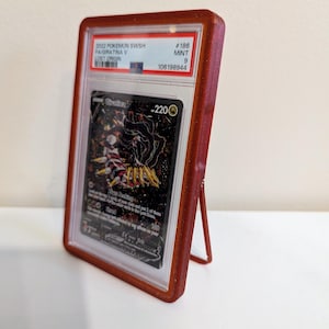 May include: A rust-colored swivel stand slab case holding a graded 2022 Pokemon SWSH Giratina V card. The case features a built-in kickstand on the back. Text on the image reads Swivel Stand Slab Case and PATENT PENDING.