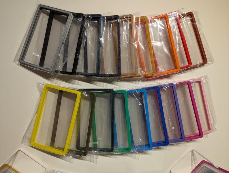 May include: A collection of colorful rectangular frames in various hues, including yellow, green, blue, and pink, are displayed. Each frame is individually packaged in clear plastic. The frames are arranged in two rows, showcasing a range of colors.